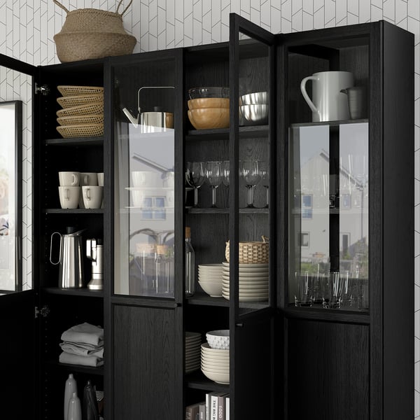 Dark wooden bookcase with glass doors, displaying cups, bowls, and wine glasses. Adjustable shelves hold woven baskets and neatly folded towels.