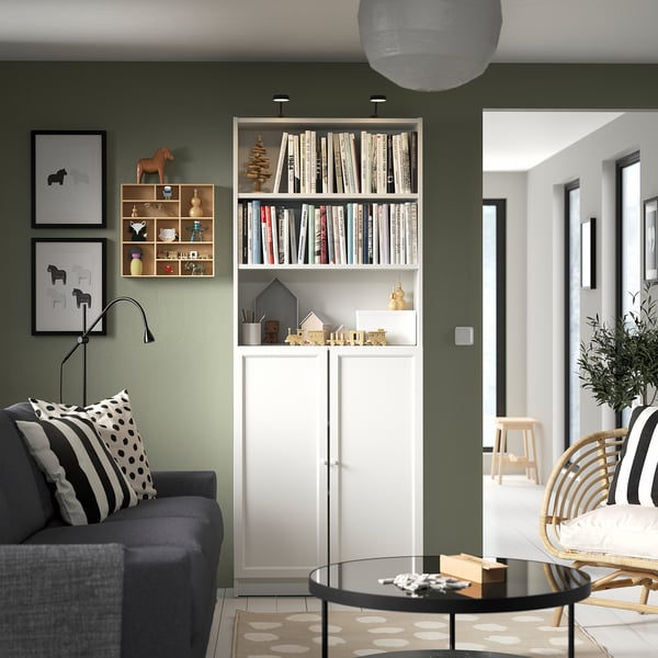 Modern living room with white BILLY bookcase against green wall, dark grey sofa, glass coffee table, and black & white accents.