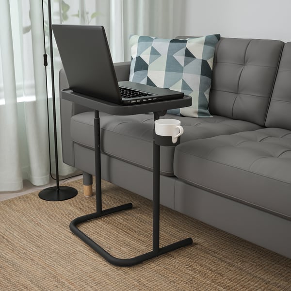 Black laptop stand with mug on steel base next to gray sofa.