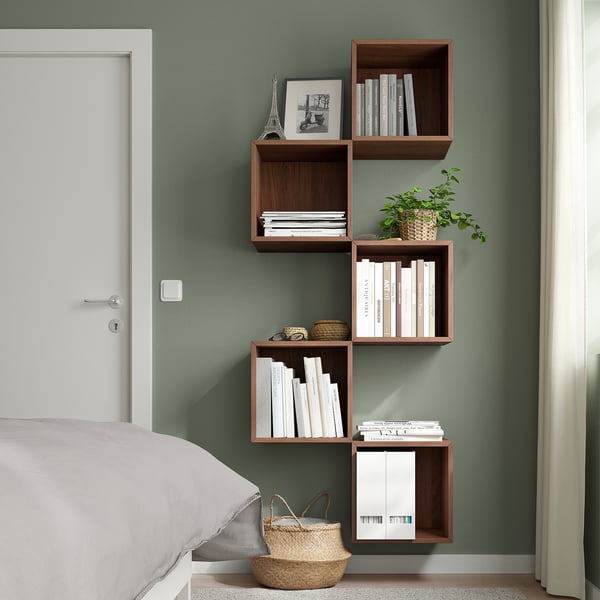 Modern bedroom with asymmetric wall-mounted dark brown EKET shelves, holding books, decor, and a plant, beside a white bed, and a closed white door.