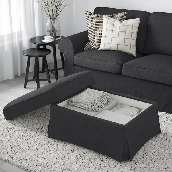 Modern living room with grey EKTORP footstool featuring storage. Sofa with black and white pillows, side table with cup and flowers.