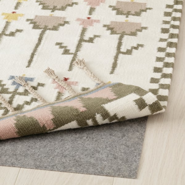 Handwoven rug with floral pattern, folded corner revealing backing and fringed edges, placed on light hardwood floor with grey rug pad underneath.