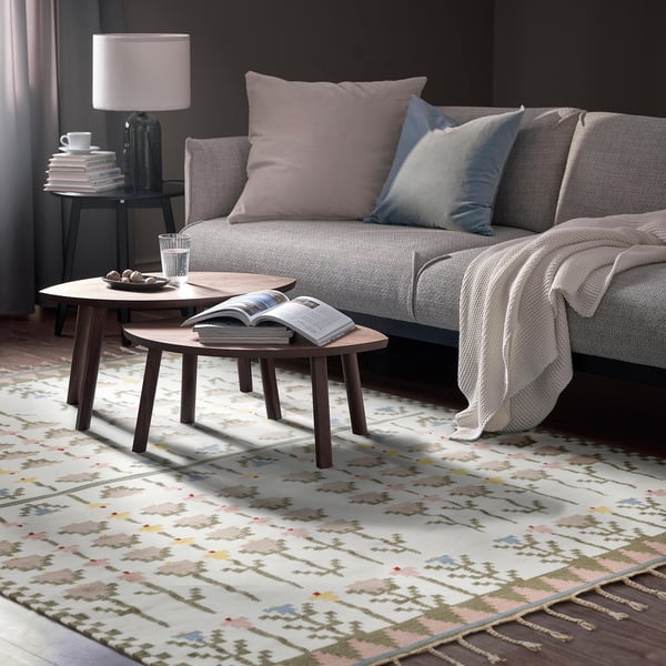 Living room scene: grey sofa with pastel pillows, two wooden coffee tables, colourful area rug, lamp with books, and blanket. Rug mainly covers floor.