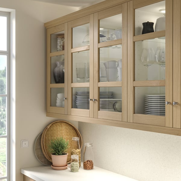 Oak kitchen cabinets with glass doors showing plates and glassware. White countertop holds tray, plant, and glass containers.