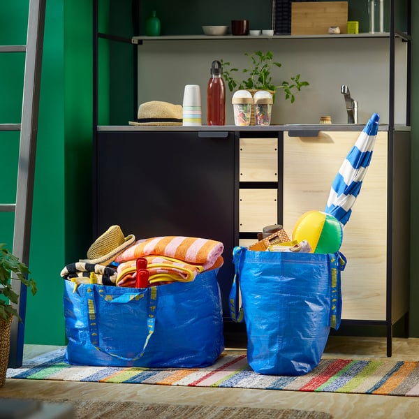Two blue FRAKTA bags filled with colourful towels and beach items are placed on a striped rug in front of a shelf with kitchen items.