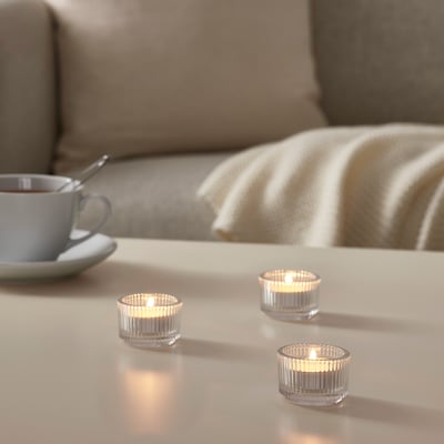 Three lit GLIMMA candles on white table, blue cup, spoon, beige blanket on couch, warm glow.