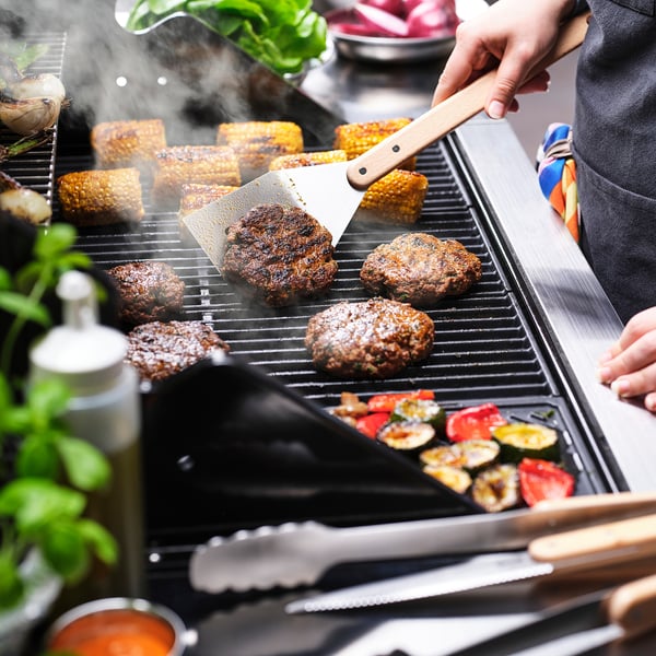 Person cooking burgers and vegetables on a grill using a spatula.