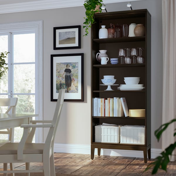 Dark brown wooden bookshelf with various items including white bowls, vases, and books; next to dining chairs and near window.