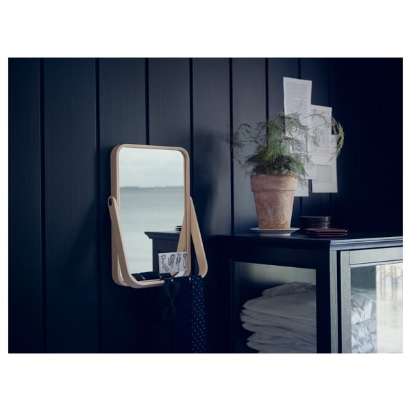 IKORNNES mirror on dark cabinet, reflecting window, with plant and folded items.