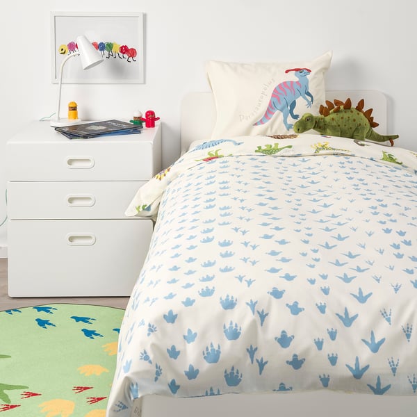 Childs dinosaur-themed room with blue bedding, green rug, dinosaur pillow, bedside table, and dresser.
