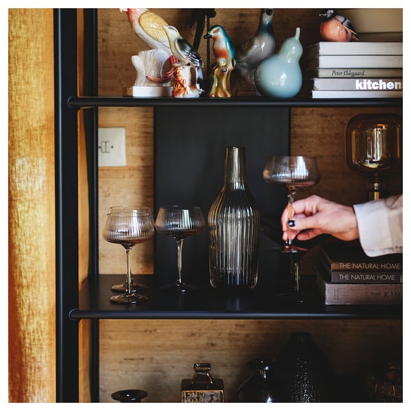 Shelf with black metal frame and bamboo shelves. Decorated with vases and figurines. A hand holding a wine glass. Books placed on upper shelf.