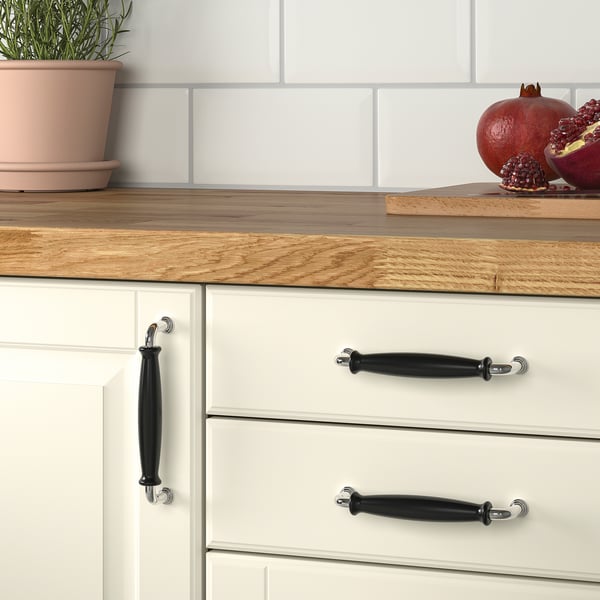 Kitchen drawers with black porcelain knobs, white cabinets, wooden countertop, potted plant, and fresh pomegranates.