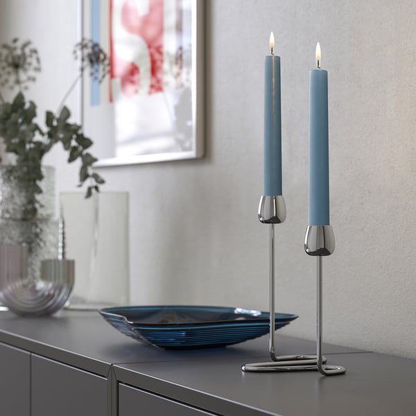 KLOKHET blue candles on metal holders, sleek, tall, set on dark furniture beside a decorative bowl.