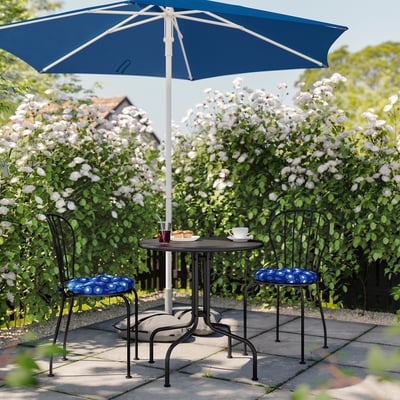 Cosy patio setup with blue umbrella, round table, and two chairs with patterned cushions surrounded by blooming white flowers.