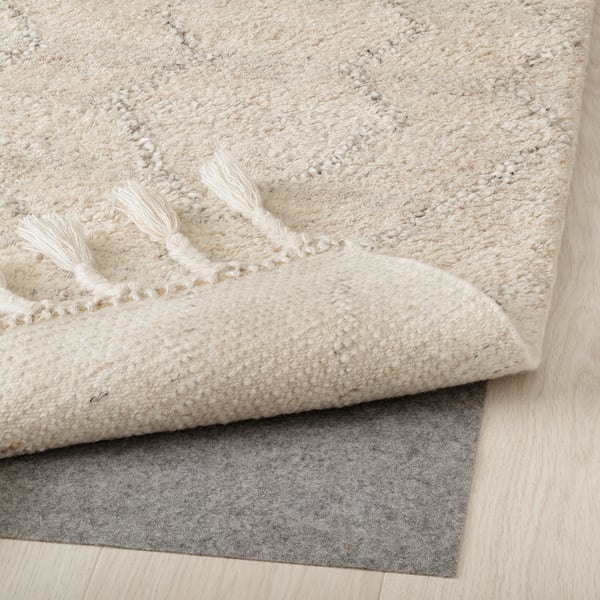 A fluffy, cream-colored wool rug with fringed edges, placed on a grey rug underlay for stability and comfort.