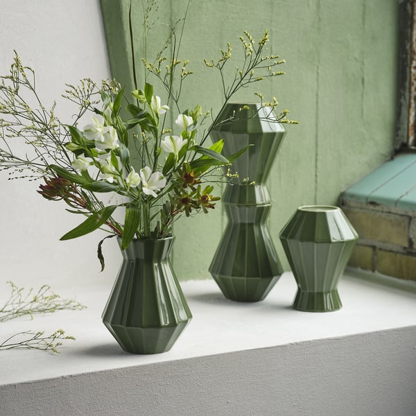 Three green geometric vases, one holding white flowers, displayed on a white surface against a green wall.