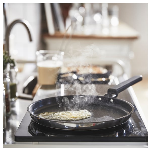 Black frying pan with steam, cooking on a stove, placed on a white countertop near a sink. A glass is nearby.