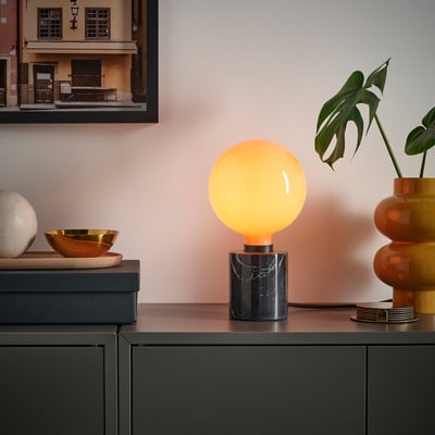 Orange MOLNART bulb in black marble base, with golden bowl, white sphere, plant, and picture.