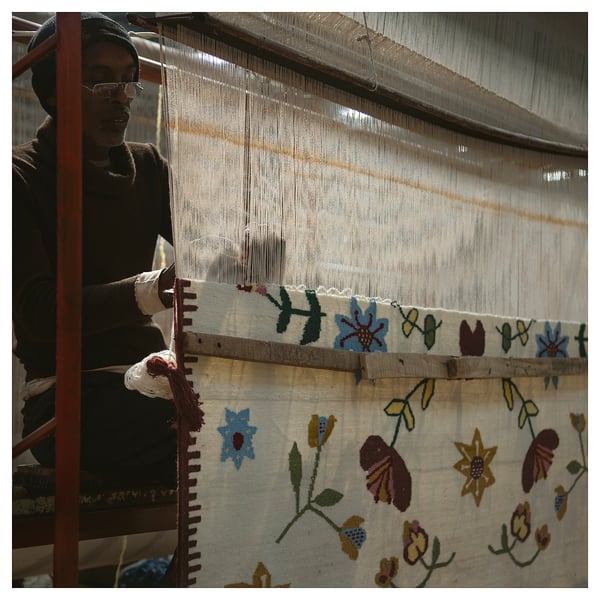 Person weaving vibrant, flower-patterned rug on loom.