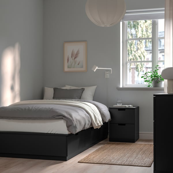 Minimalist blue bedroom with NORDLI black bed frame, white bedding, grey rug, pendant light, bedside table with plant, and window.