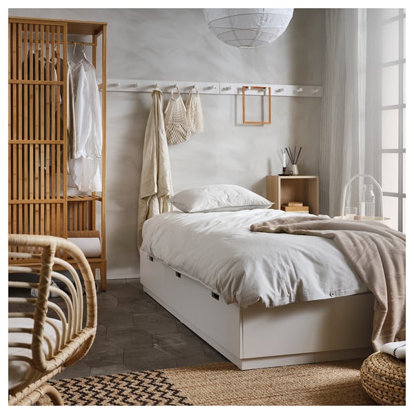 Modern minimalist bedroom with NORDLI bed, linens, brown blanket, closet, wicker chair, light grey walls, rug, decorative light.