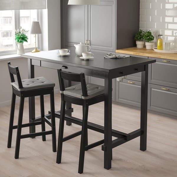 Modern kitchen with black NORDVIKEN bar table and stools, cushioned seats, near window with plants and teapot.