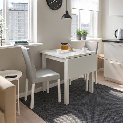 White drop-leaf table with 2 chairs in kitchen, tea set, near windows.