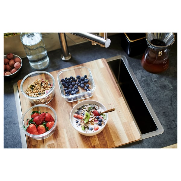 Wooden kitchen cutting board with bowls of fruit and granola on a dark countertop.