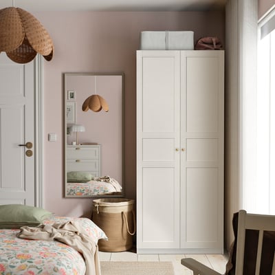 Cosy bedroom with tall white wardrobe, floral bedding, mirror, wicker basket, pendant lights, pink walls.