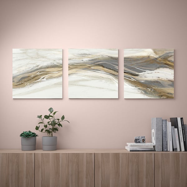 Three abstract canvas prints hang on a pink wall, flanked by potted plants and stacked books on a wooden shelf below.