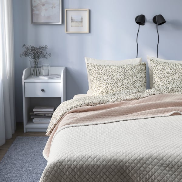 White quilted bedspread with pillows, bedside table with books and vase.