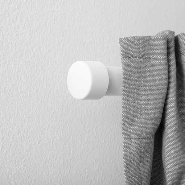 A white cylindrical curtain finial is positioned on a grey curtain rod, attaching the curtain to the wall. The finials design is clean and minimalist.