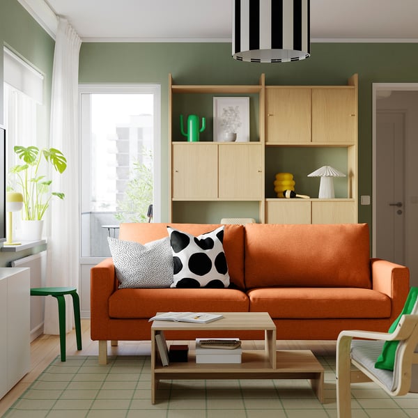 Modern living room with orange sofa, black-white pillows, wooden table, green armchair, bookshelf with decor, and large window.