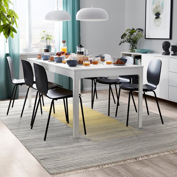 Modern dining set with white table, black chairs, striped rug, food, and pendant lamps.