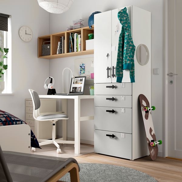 A teen room with a white SMÅSTAD wardrobe, desk, and chair. The wardrobe is tall with drawers, hooks, and a mirror. A skateboard leans against it.