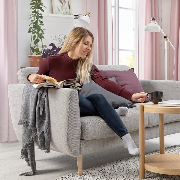 A person reads a book on a grey SMEDSTORP sofa by a window. The room is decorated with pink curtains and a plant.