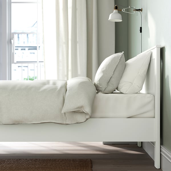 Modern bed with white striped pillows, positioned near a window with sheer curtains and a wall-mounted lamp.