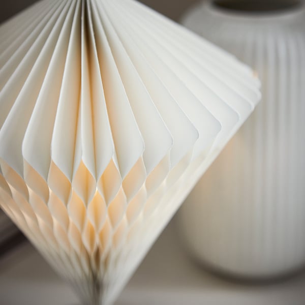 Close-up of modern, white paper lamp with intricate pleated design; soft glow, battery-powered.