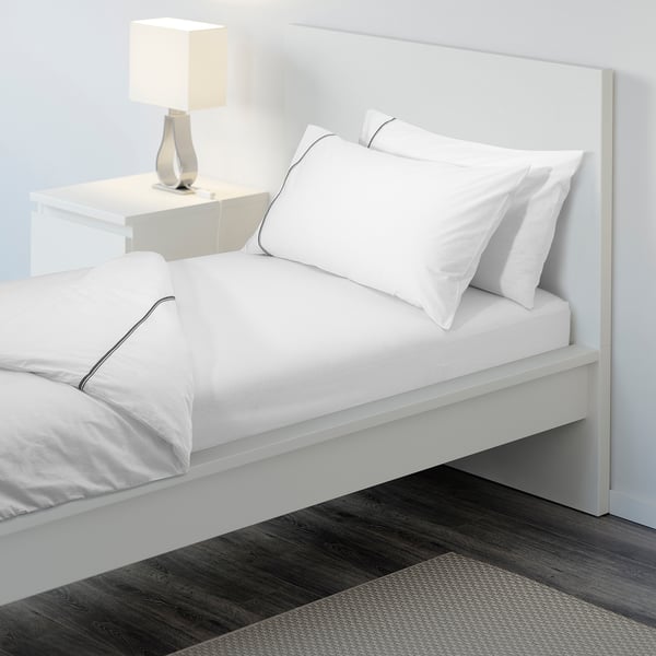 A neatly made bed with white sheets and pillows, elastic edges visible. A bedside table with a lamp beside it.