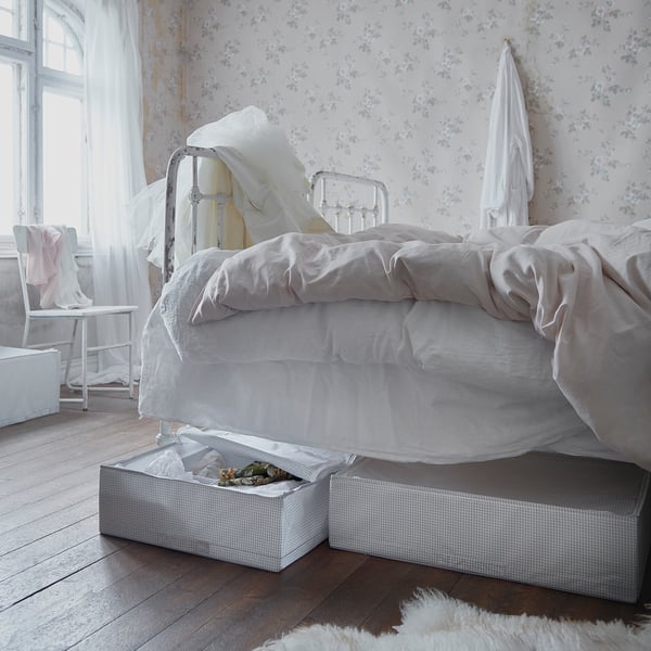 Vintage bedroom with patterned wallpaper, metal bed, white linens, and storage boxes.