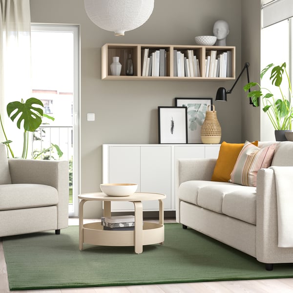 Modern living room with light-colored sofas, a central wooden coffee table, and a green wool rug that matches the plant-filled, minimalist aesthetic.