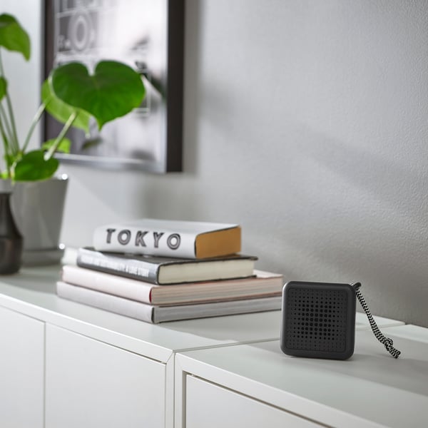 Black VAPPEBY speaker, small, square with round holes, sits on shelf by books & plant, portable, waterproof.