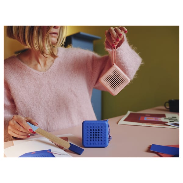 A person in a pink sweater holds a pink portable speaker while sitting at a table with craft items. A blue speaker is also on the table.