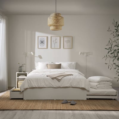 Minimalist bedroom with white VIHALS bed, light wood floor, beige rug, grey slippers, white pillows, and hanging light. Three framed photos above bed.