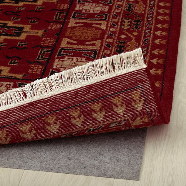 Red rug with intricate patterns and fringed edges. Grey underlay visible, providing stability. Ideal for various floors, adding warmth and texture.
