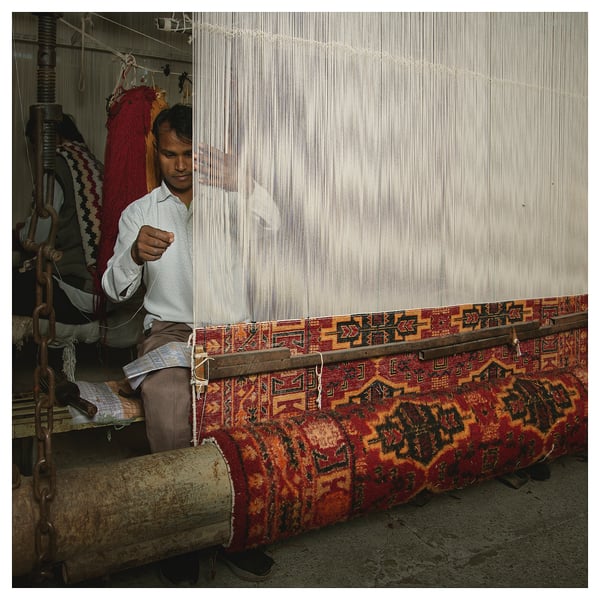 Person weaving colourful, patterned rug on traditional loom. Rug red, yellow, and black designs. Loom has white threads.