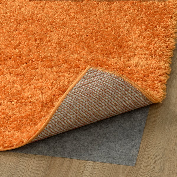 Ljusorange lurvig matta med texturerad grå halkfri dyna under, placerad på trägolv.