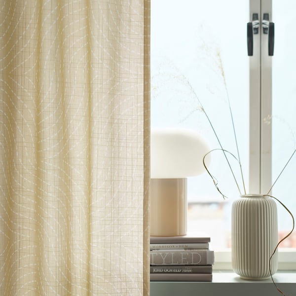 ÅKERMOLKE gardiner, beige, strukturerade, hänger nära ett fönster, bredvid en hög vas och staplade böcker.