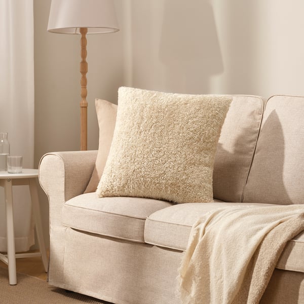 Mysig beige ÅSJORDFLY fyrkantig prydnadskudde på ljus soffa, mjuk teddypälstextur.
