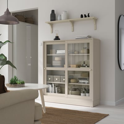 BERGSHULT beige hylla med glasdörrar, förvaring av föremål, under vägghylla, nära soffa och växtbord.